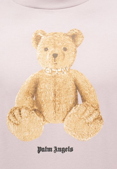 Ανοιχτό καφέ αρκουδάκι teddy με κεντημένο γιακά πάνω από το κείμενο «Palm Angels» σε φόντο από ανοιχτό ροζ ύφασμα.