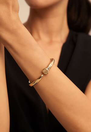 Primer plano de una persona que lleva una pulsera pulida de oro, abierta en un extremo, en la muñeca, sobre un fondo negro de camiseta.