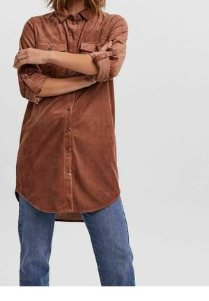 Bruine ribfluwelen blouse met knoopsluiting aan de voorkant, twee borstzakken en opgerolde mouwen. Losse pasvorm gecombineerd met blauwe jeans met rechte pijpen.