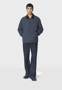 Mørkeblå quiltet jakke med knaplukning foran, stående krave, og klaplommer, båret over mørke denimbukser og sorte sko.