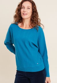 Türkiser Strickpullover mit weitem Ausschnitt und überschnittenen Schultern, ausgestattet mit gerippten Bündchen und Saum sowie einem kleinen metallischen Logo-Detail.