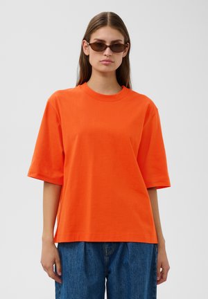 Ung kvinde iført brune solbriller, en lys orange oversized kortærmet skjorte og løse blå jeans, stående mod en grå baggrund.