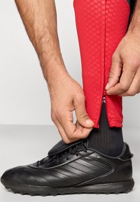 Røde træningsbukser med et tekstureret mønster og lynlåse ved anklerne, kombineret med sorte træningssko med en glat overflade og snørebånd.