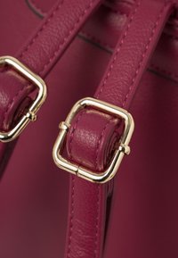 Dettaglio della tracolla in pelle magenta con hardware dorato e accenti plissettati testurizzati. I bordi cuciti mettono in risalto il design.