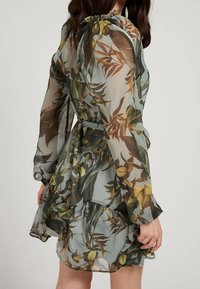 Vestido transparente de un verde claro con estampado floral en amarillo y marrón. Tiene mangas largas, escote fruncido y dobladillo con volantes en capas.