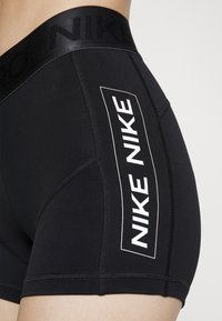 Mustat urheilushortsit, joissa on silikoninen vyötärönauha kohokuvioidulla logolla, valkoinen "NIKE"-teksti sivussa ja ommellut saumat kestävyyttä varten.