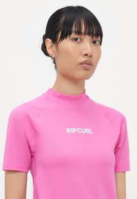 Pink short-sleeve rash guard featuring a mock neck and the "RIPCURL" logo in white across the chest. Smooth texture and fitted design.