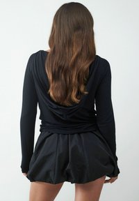 Schwarzes Langarm-Hoodie mit einer drapierten Kapuze, kombiniert mit einem schwingenden schwarzen Rock aus leichtem Material, der ausgeprägte Falten aufweist.