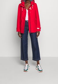 Chaqueta impermeable roja con capucha, forro interior blanco y cordón ajustable. Combinada con vaqueros anchos azules y zapatillas multicolores.