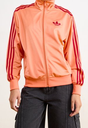 Personne portant un blouson de sport pêche avec fermeture éclair, bandes rouges et logo sur les manches, associé à un pantalon gris foncé avec poches latérales.