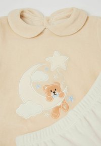 Beżowy strój niemowlęcy z haftowanym misiem na półksiężycu. Miękki materiał, okrągły kołnierzyk i elastyczne białe spodnie.