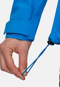 Manica di giacca impermeabile blu con polsino regolabile e cordino. Mano che afferra un cordino elastico, mostrando una texture liscia e resistente.