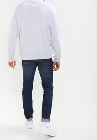 Felpa grigia con colletto e polsini a costine, abbinata a jeans blu scuro e sneakers bianche, vista da dietro su uno sfondo neutro.