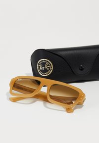 Ochelari de soare Ray-Ban cu lentile de culoare chihlimbar și o ramă galbenă lucioasă, așezați pe un capac din piele neagră cu un logo auriu.