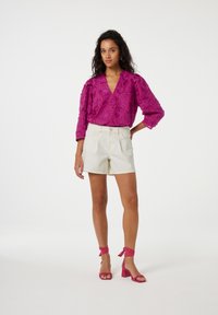 Fuchsia geborduurde blouse met pofmouwen, gecombineerd met crèmekleurige short. Roze hakken met enkelwikkeldetail maken de outfit compleet.