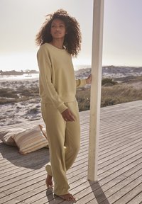 Loungewear-Set aus weichem, hellgelben Rippstoff, bestehend aus einem lässigen Oberteil und einer Hose, kombiniert mit barfuß auf einem Holzboden.