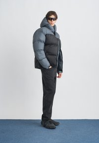 Puffer bunda v čiernej a sivej farbe, s vysokým golierom, zapínaním na zips a oversize strihom. Nosí sa s čiernymi džínsami a športovou obuvou.