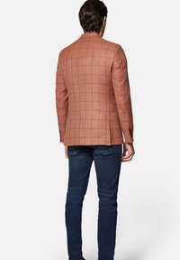 Brązowa marynarka w kratę z teksturowanej tkaniny, z klapami i trzema guzikami przy mankietach, zestawiona z ciemnoniebieskimi dżinsami.