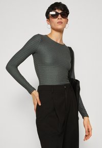 Langärmliges, strukturiertes graues Oberteil kombiniert mit hochgezogenen schwarzen Hosen. Verfügt über ein tailliertes Design und eine lässige schwarze Tragetasche.