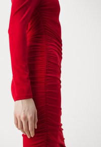 Rochie roșie dintr-un material elastic, cu un detaliu de pliere verticală pe laturi și un design cu mâneci lungi. Textură netedă, formă ajustată.