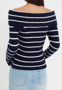 Marineblauwe en witte gestreepte off-the-shoulder trui met ribbelstructuur en fitted ontwerp. Lange mouwen en een cropped lengte benadrukken de vorm.
