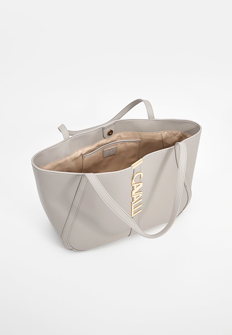 Hellgraue Tote-Tasche aus glattem Leder, mit langen Riemen und einer goldfarbenen Markenverzierung auf der Vorderseite. Offenes Innenfutter zeigt den Stoff.