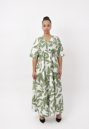 Femme portant une robe maxi blanche avec un imprimé de feuilles vertes et des manches courtes ballonnées, debout devant un fond blanc uni.