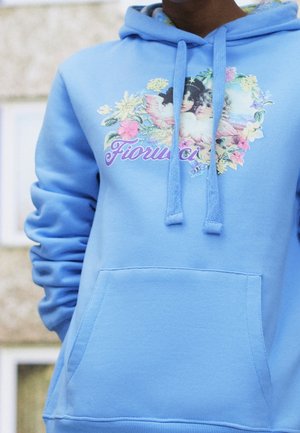 Personne portant un sweat à capuche bleu clair avec un motif floral, deux chérubins et le mot "Fiorucci" sur la poitrine.