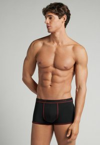 Bañadores negros con acentos rojos, hechos de una tela elástica. Tienen un ajuste ceñido y una cinturilla elástica etiquetada como "HUMAN". Textura suave.
