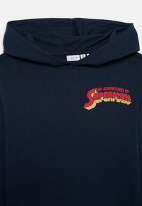 Sudadera azul marino con un gráfico frontal que dice "Las Aventuras de Superman" en rojo y amarillo, con un diseño de fuente clásica.