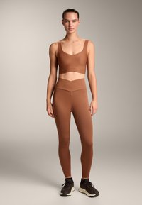 Barna, varrás nélküli sportmelltartó és magas derekú leggings, mindkettő rugalmas anyagból készült. A cropped kialakítás jellemzője egy kontrasztos ejtőszalag a deréknál.