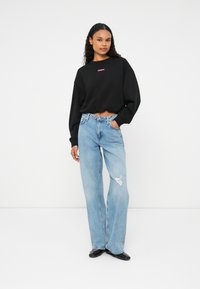 Sweatshirt negru scurt, cu un logo roz și o croială lejeră, asortat cu jeansi albaștri deschiși, cu talie înaltă și picioare largi, având detalii uzate.