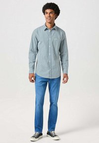 Zielono-biała koszula w kratę z długim rękawem, zestawiona z niebieskimi dżinsami i czarnymi trampkami. Koszula ma detal w postaci kieszonki.