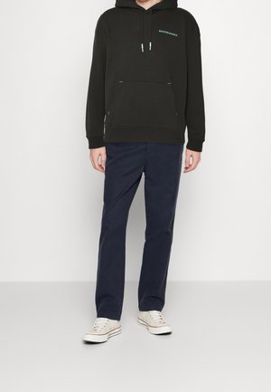 Svart huvtröja med en framficka, med turkosa detaljer; matchad med marinblå byxor och benvita sneakers.