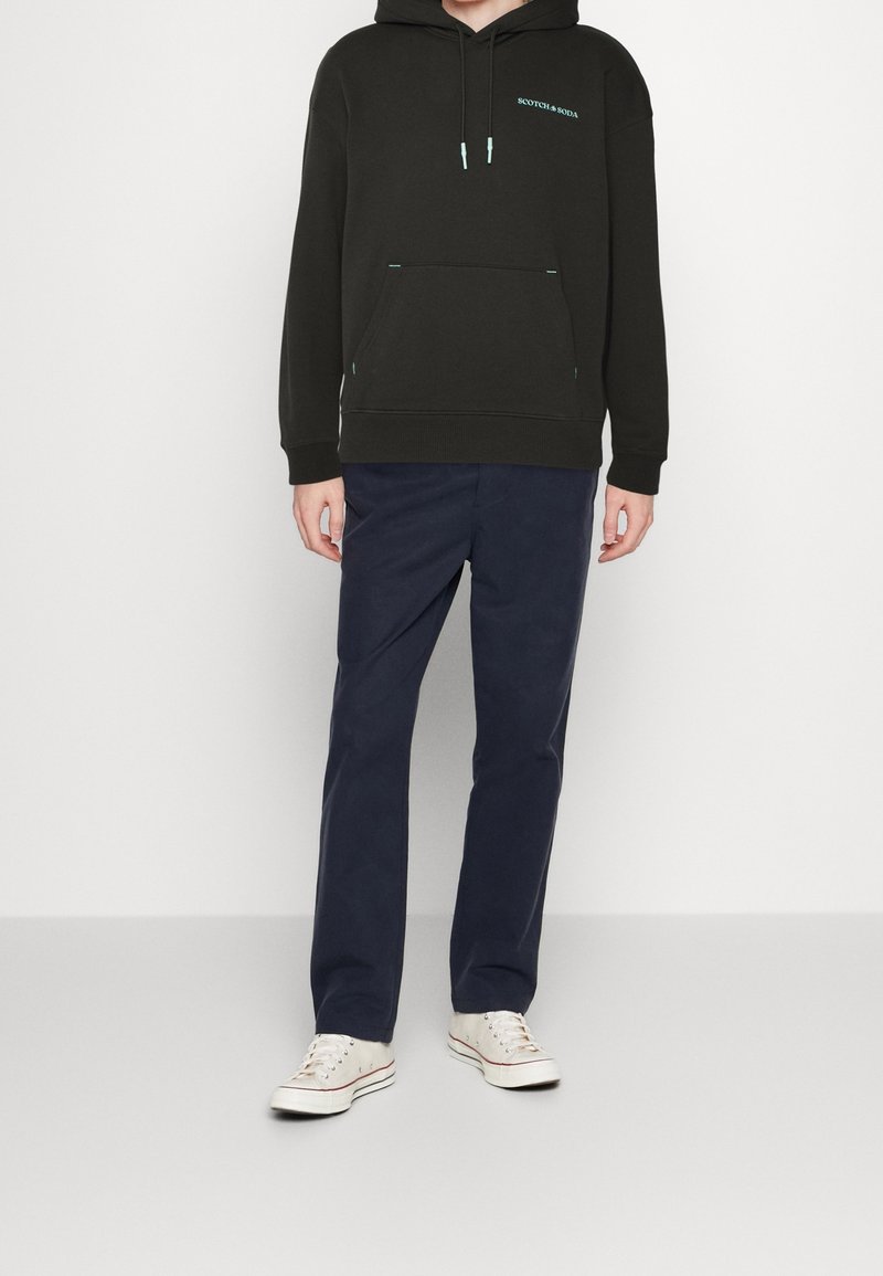 Sudadera negra con capucha y un bolsillo frontal, con acentos turquesa; combinada con pantalones azul marino y zapatillas color beige.