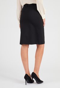 Falda lápiz negra con cintura elástica, hasta la rodilla, de textura suave, combinada con zapatos de tacón altos negros, vista desde atrás.
