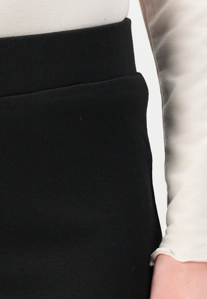 Gonna nera elasticizzata con una texture liscia e una vita aderente. Un top a maniche lunghe di colore chiaro parzialmente visibile sulla destra.