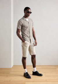 Camisa de manga curta com um padrão geom�étrico castanho, combinada com calções bege e sapatos pretos. O modelo usa óculos de sol pretos e meias brancas.