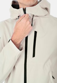 Giubbotto impermeabile beige chiaro con cappuccio, con cerniere e dettagli neri, dalla texture liscia e cordini regolabili.