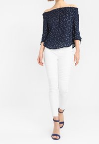 Donna che indossa un top Navy con spalle scoperte e piccoli motivi bianchi, pantaloni bianchi slim-fit e sandali aperti Navy, che cammina in avanti su uno sfondo bianco.