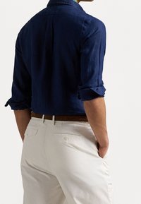 Laivastonsininen pellavapaita, jossa on käänteet hihoissa, takapuolella laskos ja kaulus. Yhdistetty vaaleanbeigeihin housuihin, joissa on takataskut ja ruskea vyö.