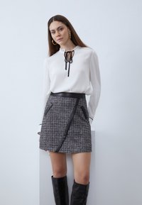 Biela blúzka s čiernou kravatu ako detail, kombinovaná s textúrovanou čierno-sivou tweedovou sukňou s koženými akcentmi a asymetrickým strihom.