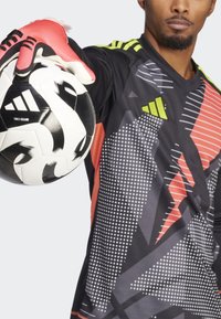 Zwarte doelman jersey met geometrische patronen in grijs, rood en neon groen accenten. Handen met rode handschoenen houden een zwart-wit voetbal vast.
