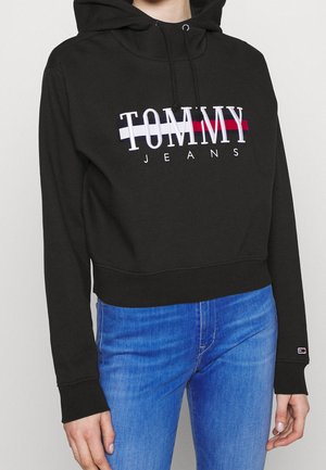 Personne portant un sweat à capuche noir avec le texte "Tommy Jeans" et des rayures rouges, blanches et bleues, associé à un jean bleu en denim.