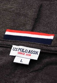 Dunkelgrauer Stoff mit einem U.S. Polo Assn. Etikett, das die Größe L zeigt, und einem rot-weiß-blauen Streifenmuster.