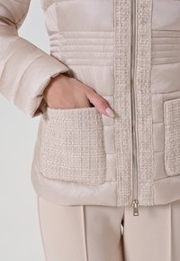 Veste beige rembourrée avec une bordure en tissu texturé, dotée d'une fermeture éclair et de deux poches avant. Surface lisse avec un éclat subtil.