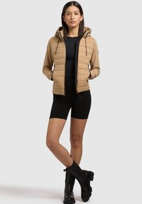 Beige polstret jakke med hætte, lynlås og snoretræk. Sort tætsiddende top og shorts med knælængde. Sorte ankelstøvler med en chunky sål.