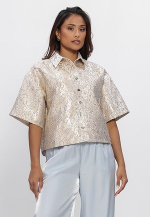 Vrouw die een beige, gestructureerde, kortärmelige blouse met knopen en lichtblauwe satijnen wijde broek draagt tegen een eenvoudige achtergrond.