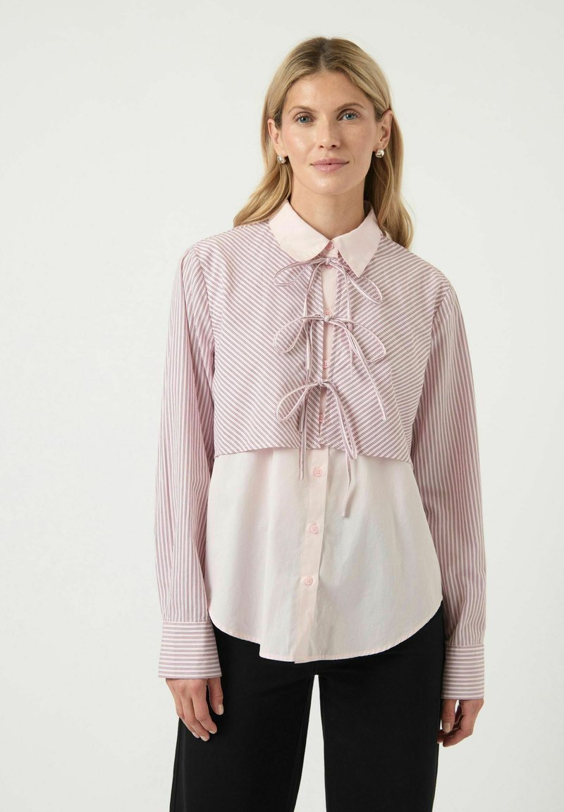 Skjorta med knäppning, med en ljusrosa och vit randig övre del, matchande slipsdetalj och en enhetlig ljusrosa nedre del. Långa ärmar.