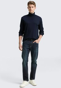 Marineblau gerippter Rollkragenpullover kombiniert mit dunkelblauen Jeans und weißen Sneakers. Das Outfit hat eine lässige Passform mit minimalen Details.